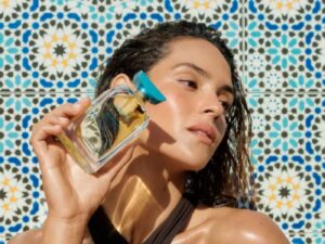 Woman in a black halterneck top holds a perfume bottle with a turquoise blue lid with Moroccanoil written on it, standing against a bright blue and yellow mosaic tiled wall.