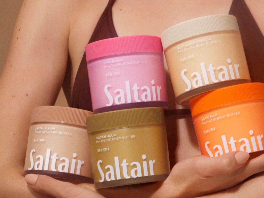 Close-up of woman in brown swimsuit holding five, different coloured pots of body care with the name Saltair on each