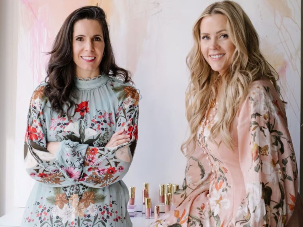 Lauren Bloomer and Natalie Mackey pictured standing side by side in an office, wearing colourful floral dresses, with lip makeup products on the desk beside them