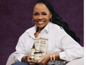 Lisa Price, wearing a white shirt and jeans and gold bracelets, seated holding a handful of Carol's Daughter haircare products.