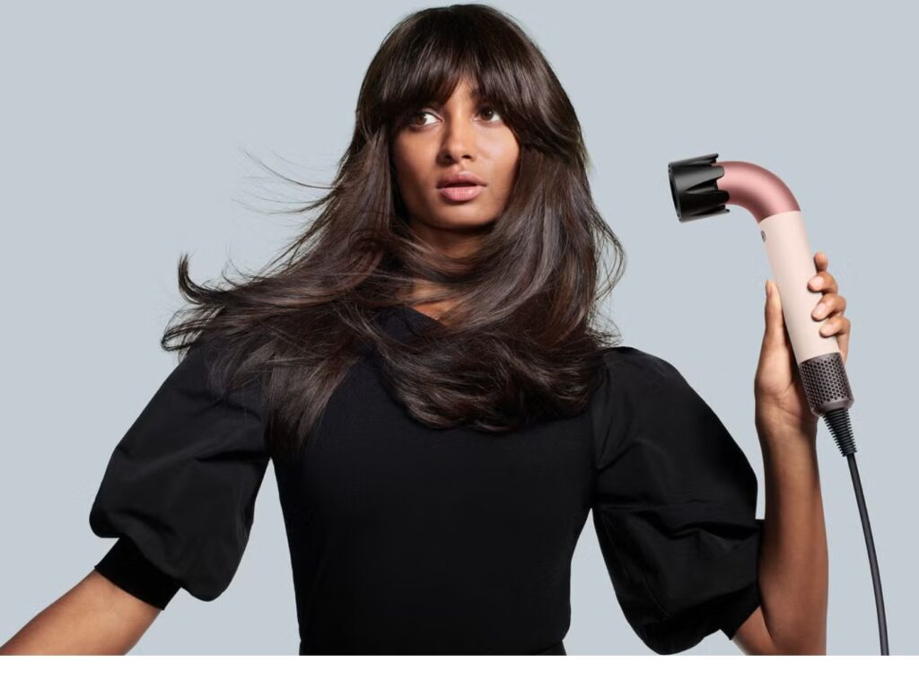 Woman with long dark hair wearing a black, puffy-sleeved sweater, holding a Dyson r, r-shaped hairdryer in rose gold, blow-drying her hair.