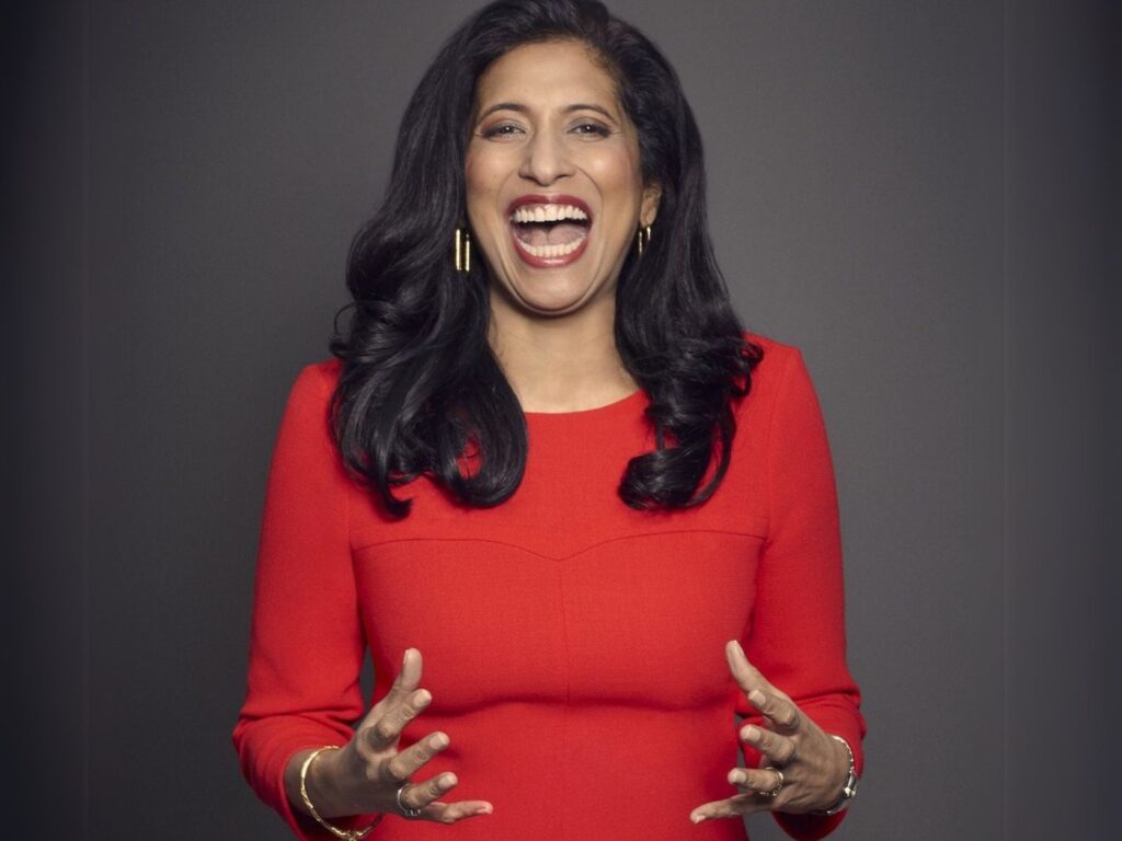 Portrait of Leena Nair wearing a red dress with three quarter length sleeves, laughing against a grey background
