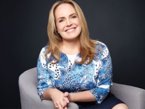 Portrait of Tara Eustace smiling, seated on a grey chair, wearing a blue print top.