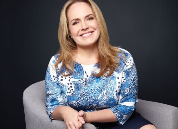 Portrait of Tara Eustace smiling, seated on a grey chair, wearing a blue print top.