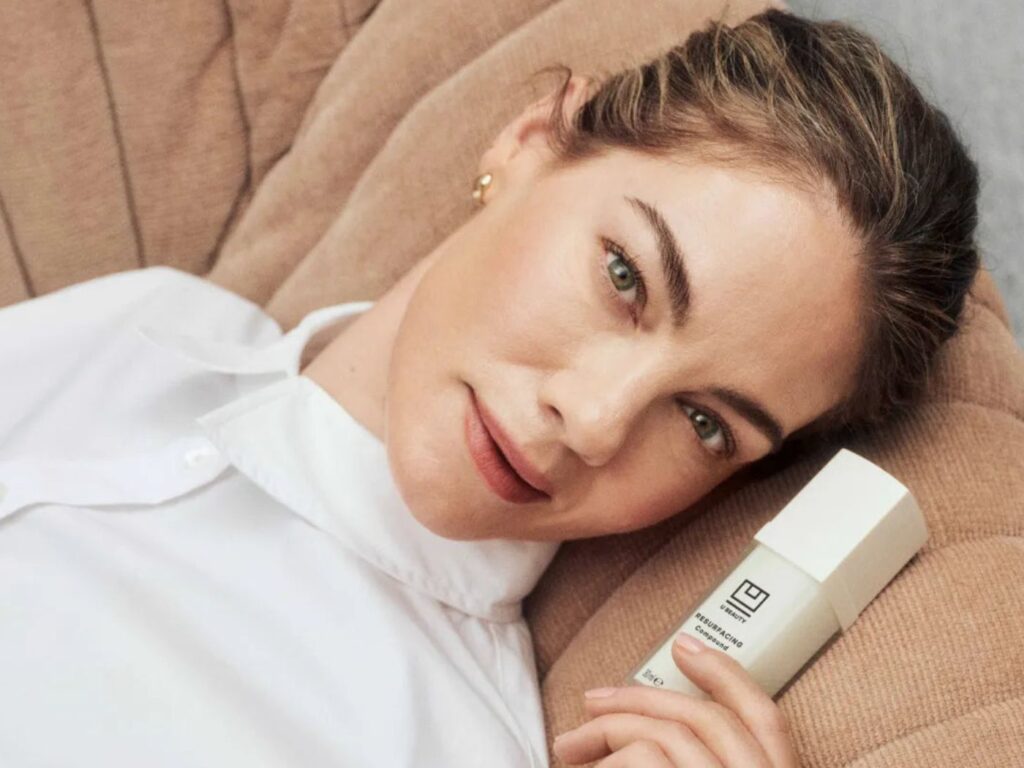 Michelle Monaghan reclining on a beige couch, wearing a white shirt, holding a bottle of beauty product with the logo U Beauty on it