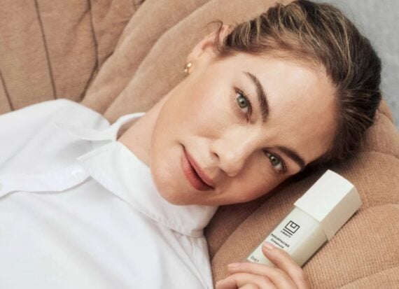 Michelle Monaghan reclining on a beige couch, wearing a white shirt, holding a bottle of beauty product with the logo U Beauty on it