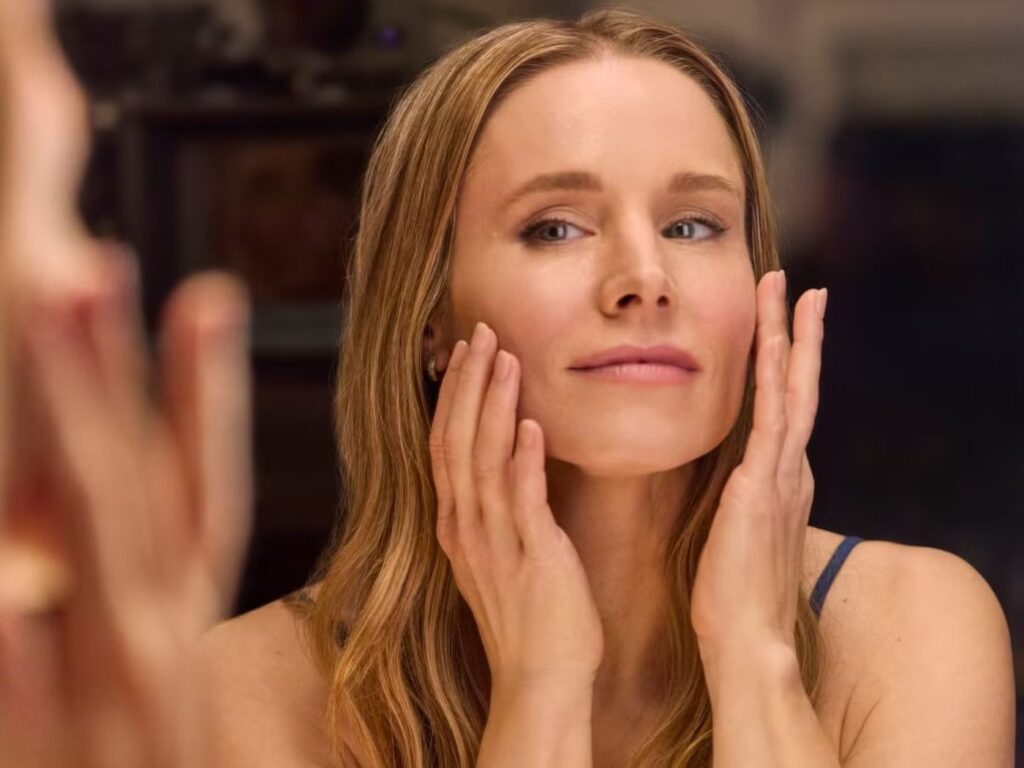 Kristen Bell looks into a mirror, doing her evening skin care routine, touching her face with both hands