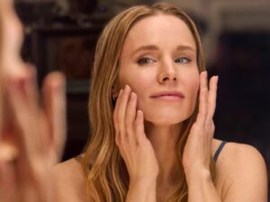 Kristen Bell looks into a mirror, doing her evening skin care routine, touching her face with both hands