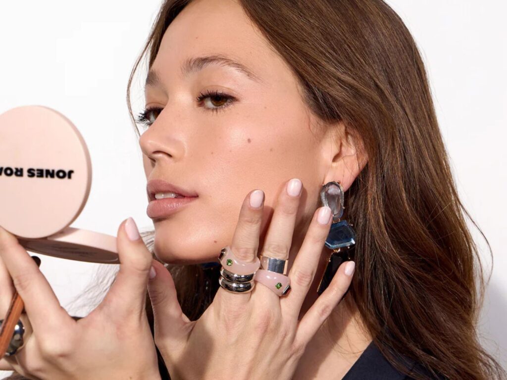 Model wearing lots of rings applies makeup to face looking in a the mirror of a pink and black Jones Road logo compact