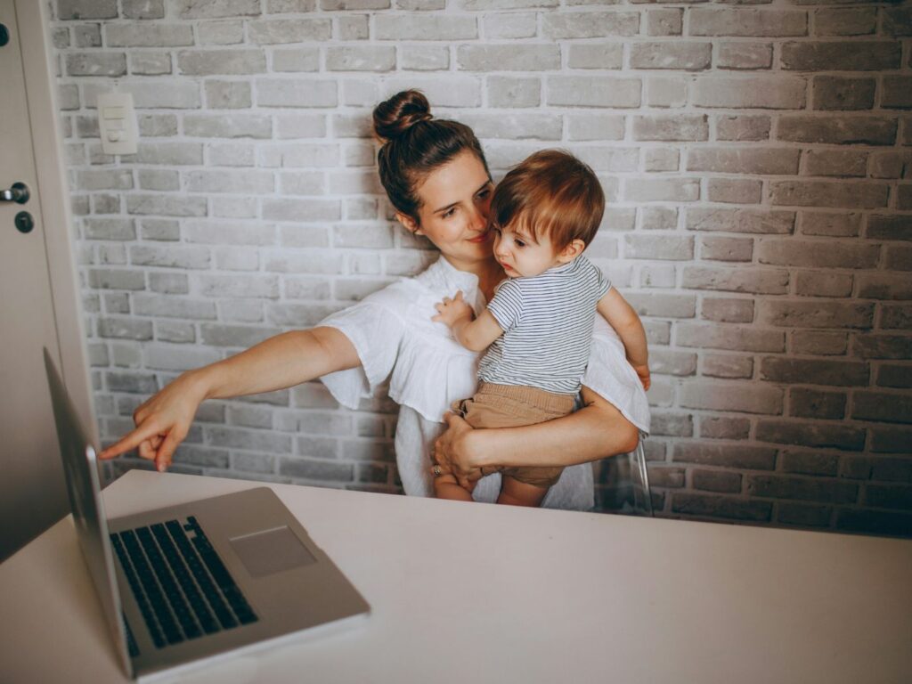 A woman seated at a large white desk at home, with her toddler son on her lap, pointing at the screen of a laptop opened on the table.