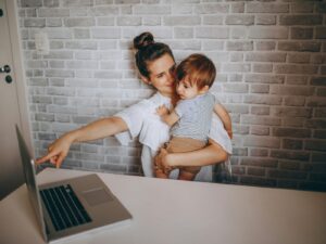 A woman seated at a large white desk at home, with her toddler son on her lap, pointing at the screen of a laptop opened on the table.