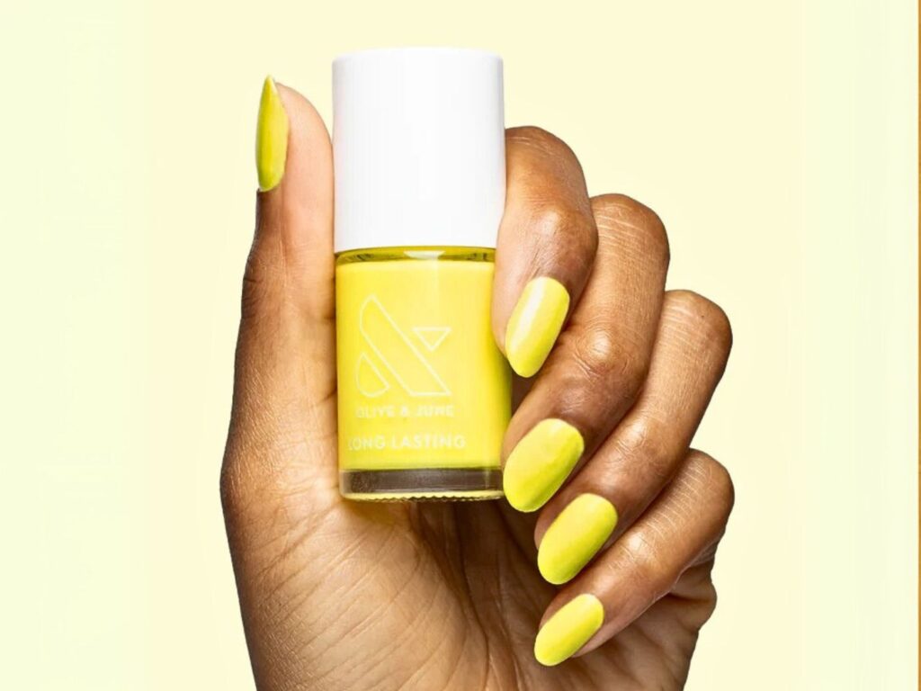 Hand with neon yellow nails holding an Olive & June nail polish bottle in the same neon yellow colour