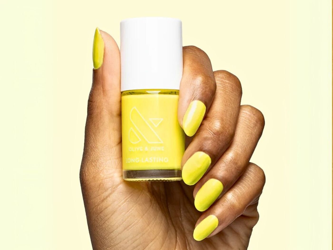Hand with neon yellow nails holding an Olive & June nail polish bottle in the same neon yellow colour