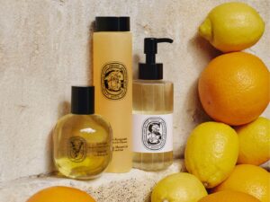 Three bottles of Diptyque bath and body products, displayed on a sand coloured stone shelf, surrounded by oranges and lemons.