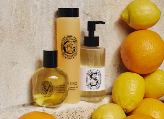 Three bottles of Diptyque bath and body products, displayed on a sand coloured stone shelf, surrounded by oranges and lemons.