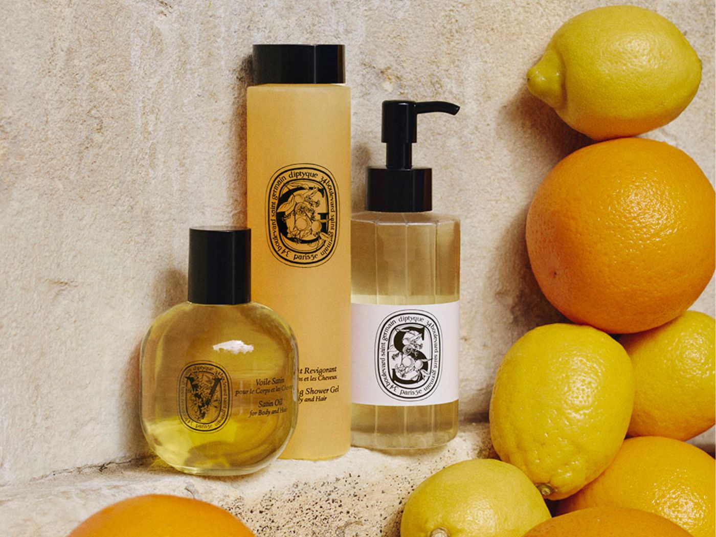 Three bottles of Diptyque bath and body products, displayed on a sand coloured stone shelf, surrounded by oranges and lemons.