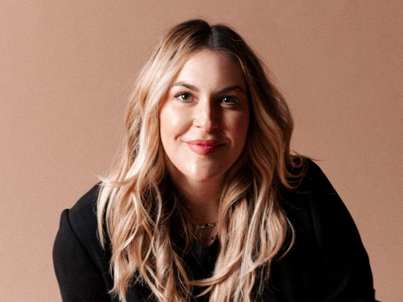 Woman with long blonde, wavy hair wearing a black sweater smiling against a peach coloured background.