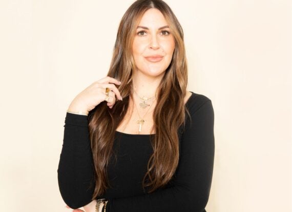 Woman with long dark blonde hair wearing a long sleeved black sweater, and layered gold necklaces and gold rings, smiling against a pale coloured background.