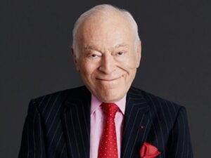 Portrait of Leonard A. Lauder, wearing a suit and pale pink shirt, with a red tie with white spots, and a red handkerchief visible in his jacket pocket, smiling.