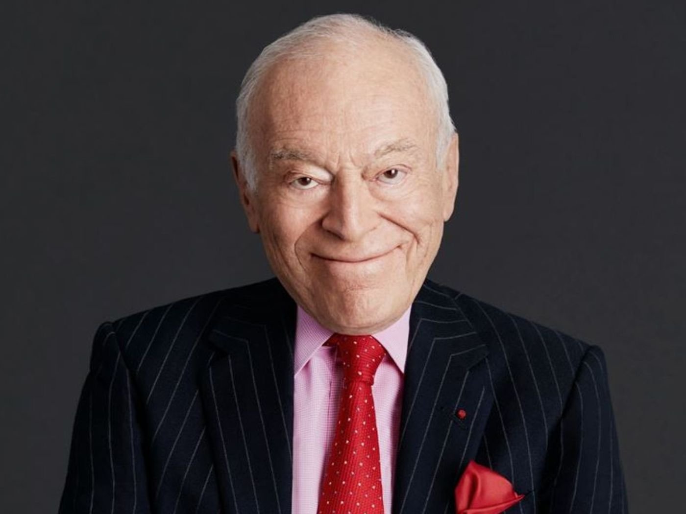Portrait of Leonard A. Lauder, wearing a suit and pale pink shirt, with a red tie with white spots, and a red handkerchief visible in his jacket pocket, smiling.