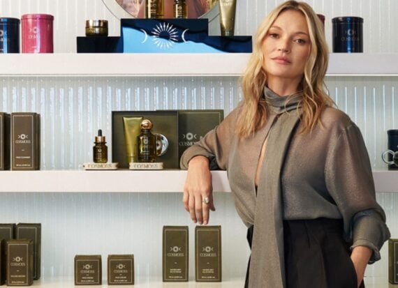 Kate Moss standing in front of shelves of Cosmoss beauty products in green and gold packaging.