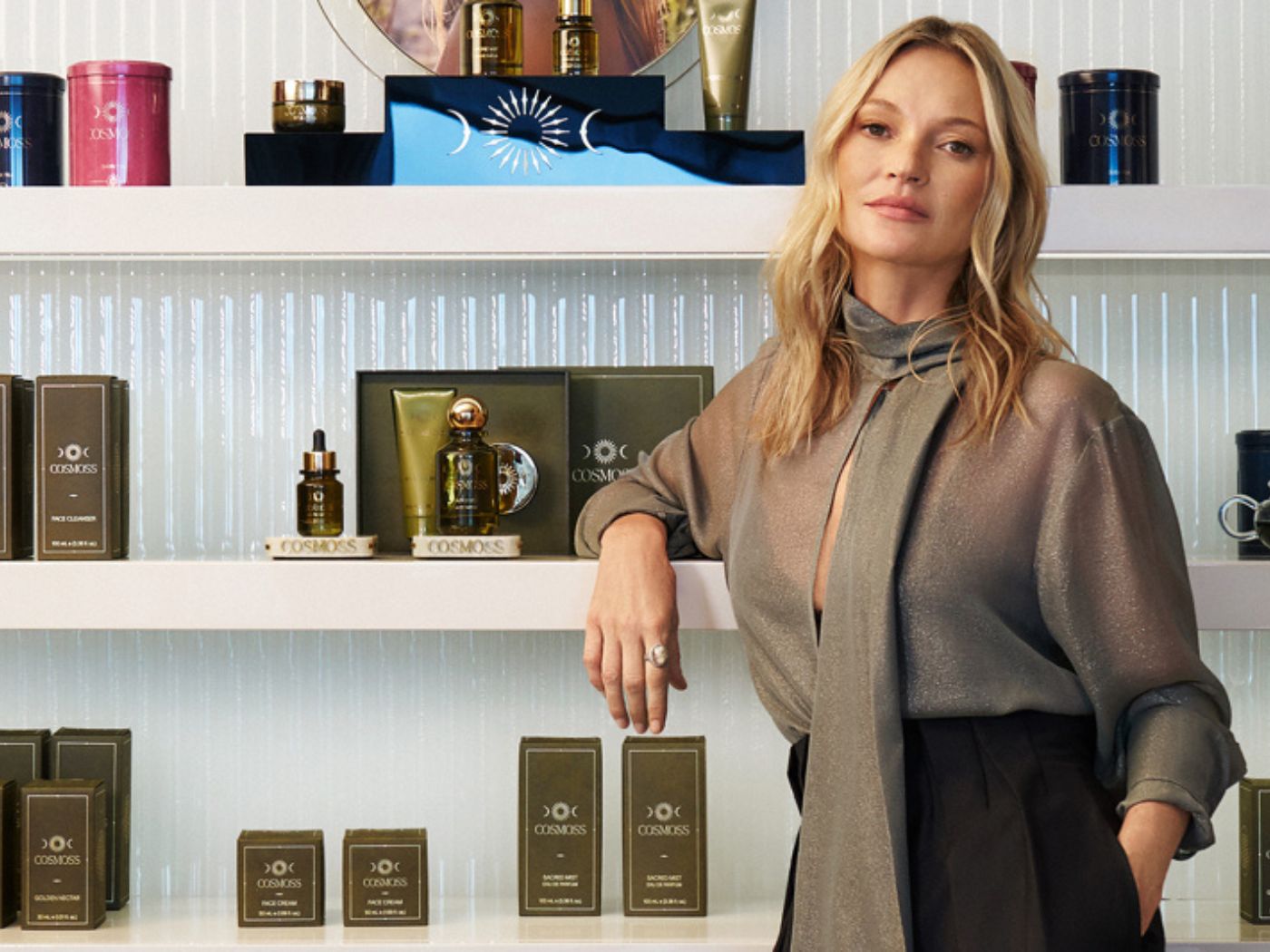 Kate Moss standing in front of shelves of Cosmoss beauty products in green and gold packaging.