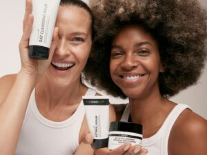 Two models, wearing white sleeveless vests, laughing holding tubes and pots of The Inkey List skin care products, in black and white packaging