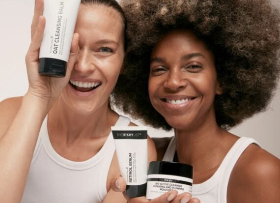 Two models, wearing white sleeveless vests, laughing holding tubes and pots of The Inkey List skin care products, in black and white packaging