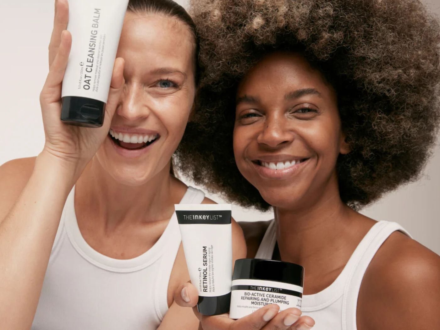 Two models, wearing white sleeveless vests, laughing holding tubes and pots of The Inkey List skin care products, in black and white packaging