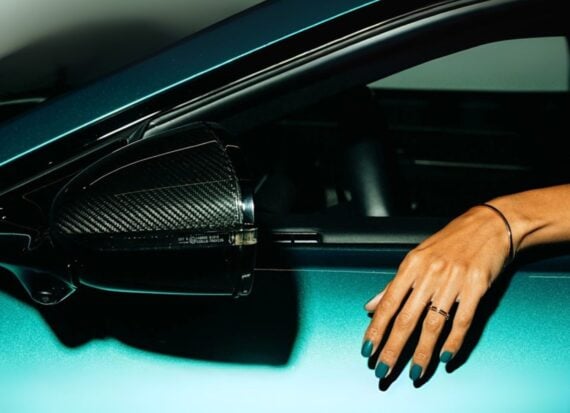 Hand with teal green manicured nails rests on the open window of an Aston Martin sports car
