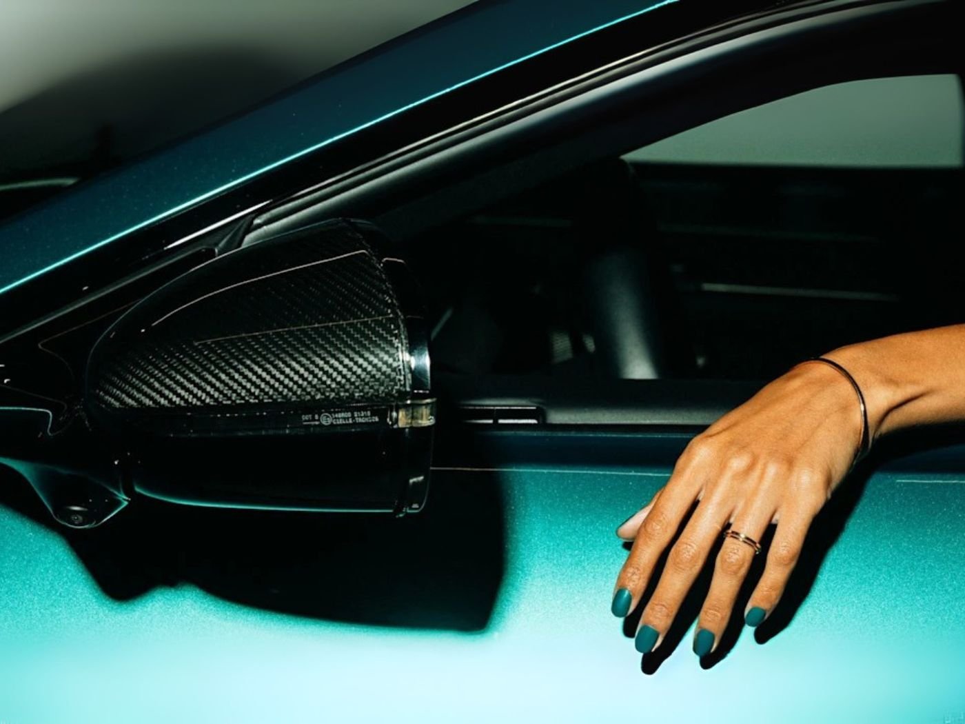 Hand with teal green manicured nails rests on the open window of an Aston Martin sports car