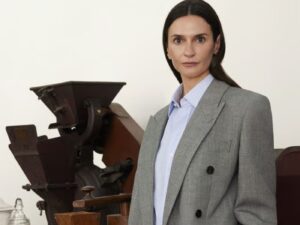 Woman with long brunette hair, wearing a grey blazer and a pale white collared shirt, stands in front of antique-looking mahogany furniture and apothecary style glass perfume bottles