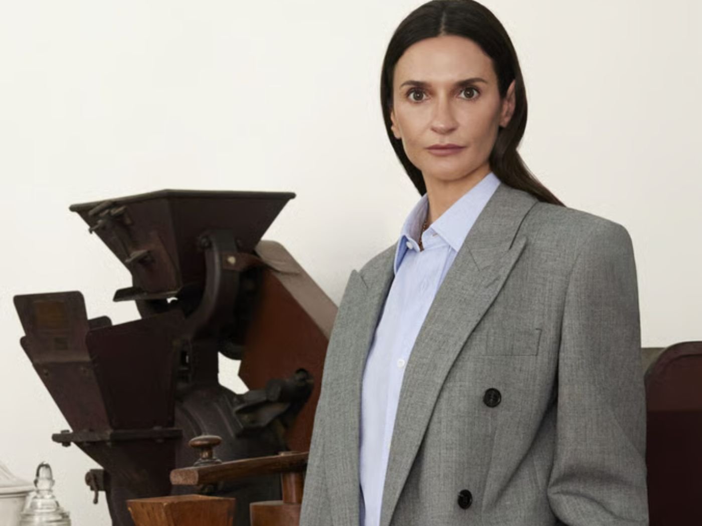 Woman with long brunette hair, wearing a grey blazer and a pale white collared shirt, stands in front of antique-looking mahogany furniture and apothecary style glass perfume bottles