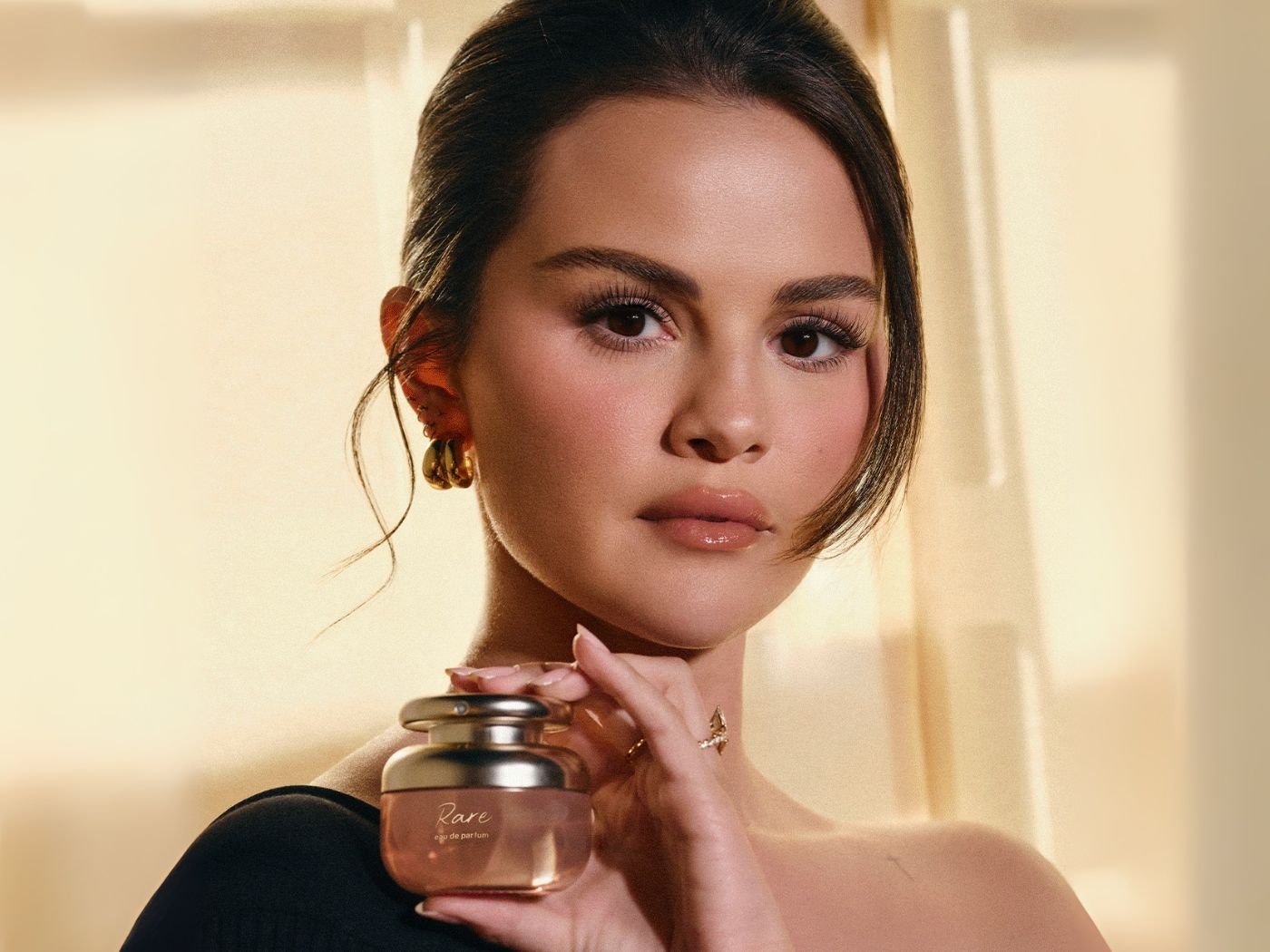 Selena Gomez in off the shoulder black evening dress and hair up holding a bottle of Rare Beauty fragrance