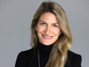 Woman with long blonde hair wearing a black turtleneck sweater and a gold necklace, smiling.