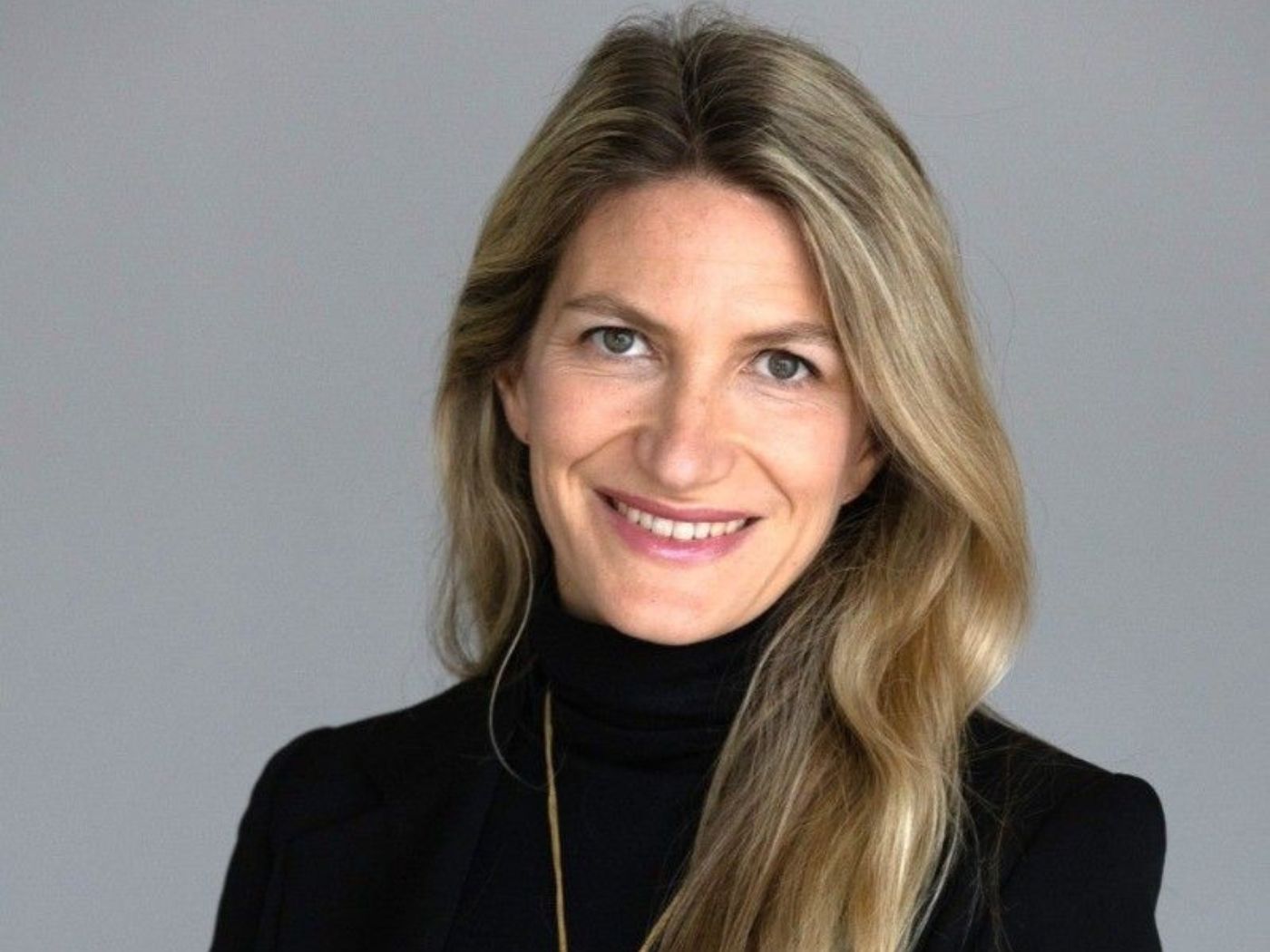 Woman with long blonde hair wearing a black turtleneck sweater and a gold necklace, smiling.