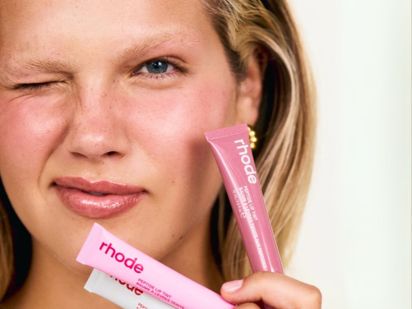 Close up of model holding a pink, cream and apricot colored tube of "rhode" lip gloss, winking at the camera