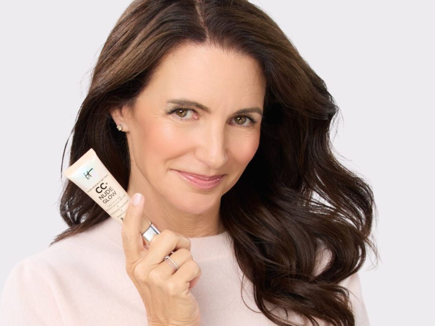 Actress Kristin Davis in a white sweater, holding a tube of IT Cosmetics makeup in one hand, smiling