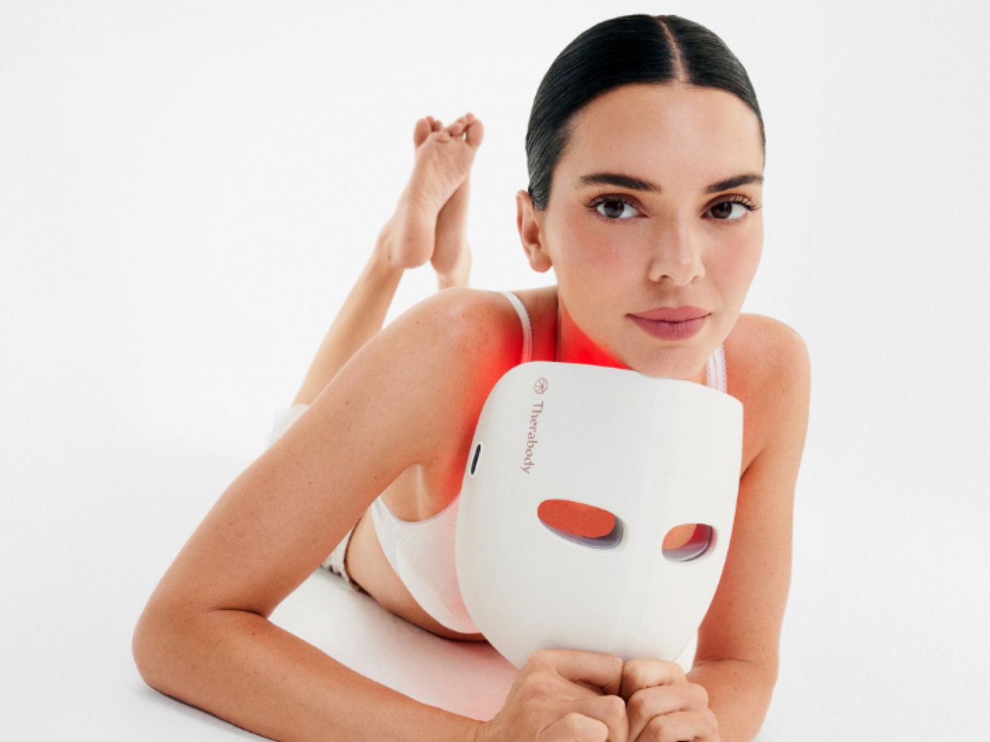 Kendall Jenner lays on a white studio floor holding a white RED LED mask in her hands