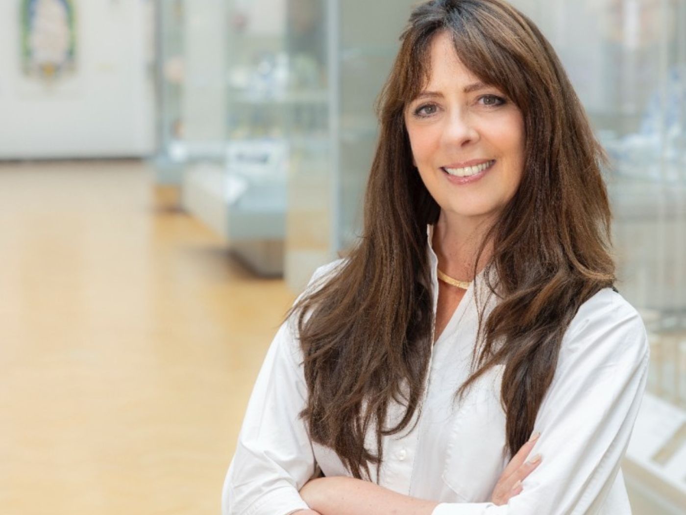 Woman with long, wavy brunette hair smiling, wearing a white shirt and a gold necklace, folding her arms.