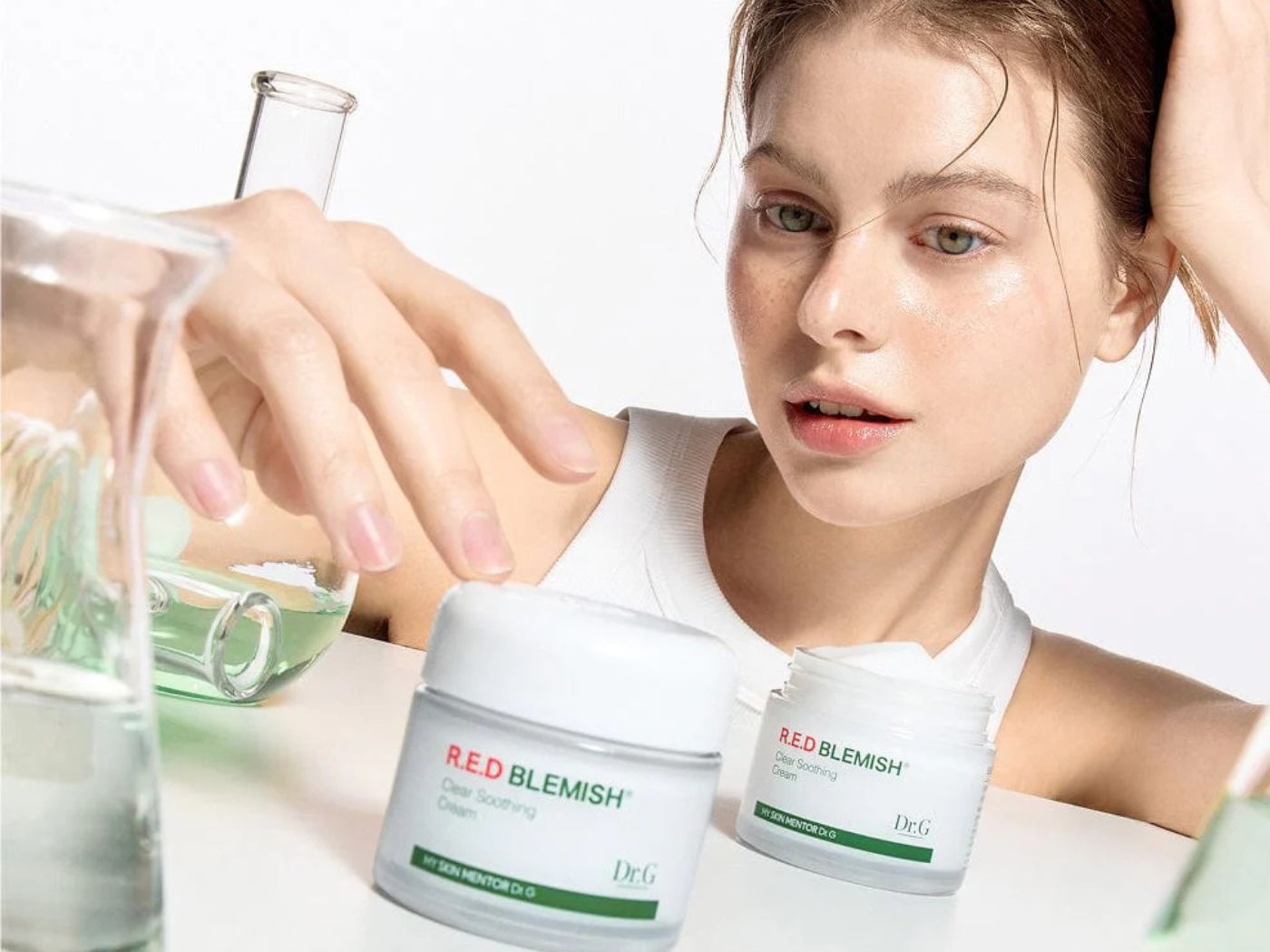 A young girl wearing a white vest top sitting om front of a white table with labaratory-style bottles of green liquid and two pots of Dr G. skin care products.