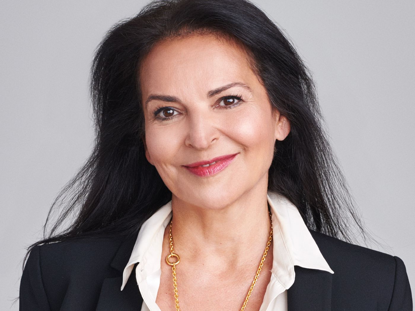 Head and shoulders photo of a woman smiling, with long dark hair wearing a white blouse and a black blazer, and a gold chain necklace.