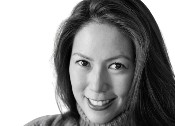 Mia Young Black and white head and shoulders photo of a woman with long dark hair smiling, wearing chunky rollneck sweater.