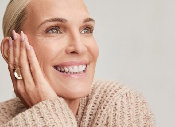 Molly Sims’ Yse Beauty Photo of a blonde woman, with hair smoothed back, smiling, wearing a gold ring and a beige chunky sweater.