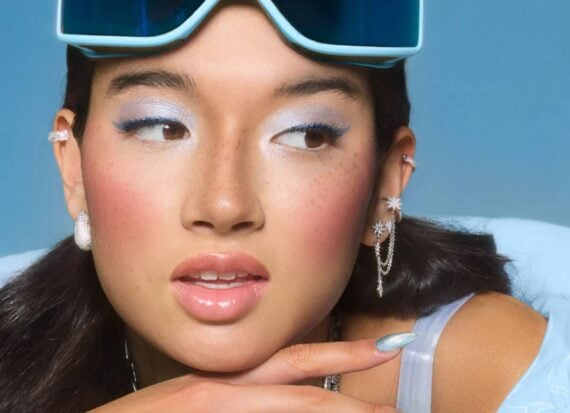 Urban Decay Head and shoulders photo of a woman with dark brown/black hair, wearing blue shimmery eye makeup and blush wearing pale blue ski goggles on her head, and with shimmering blue long nails, and a shiny blue top.